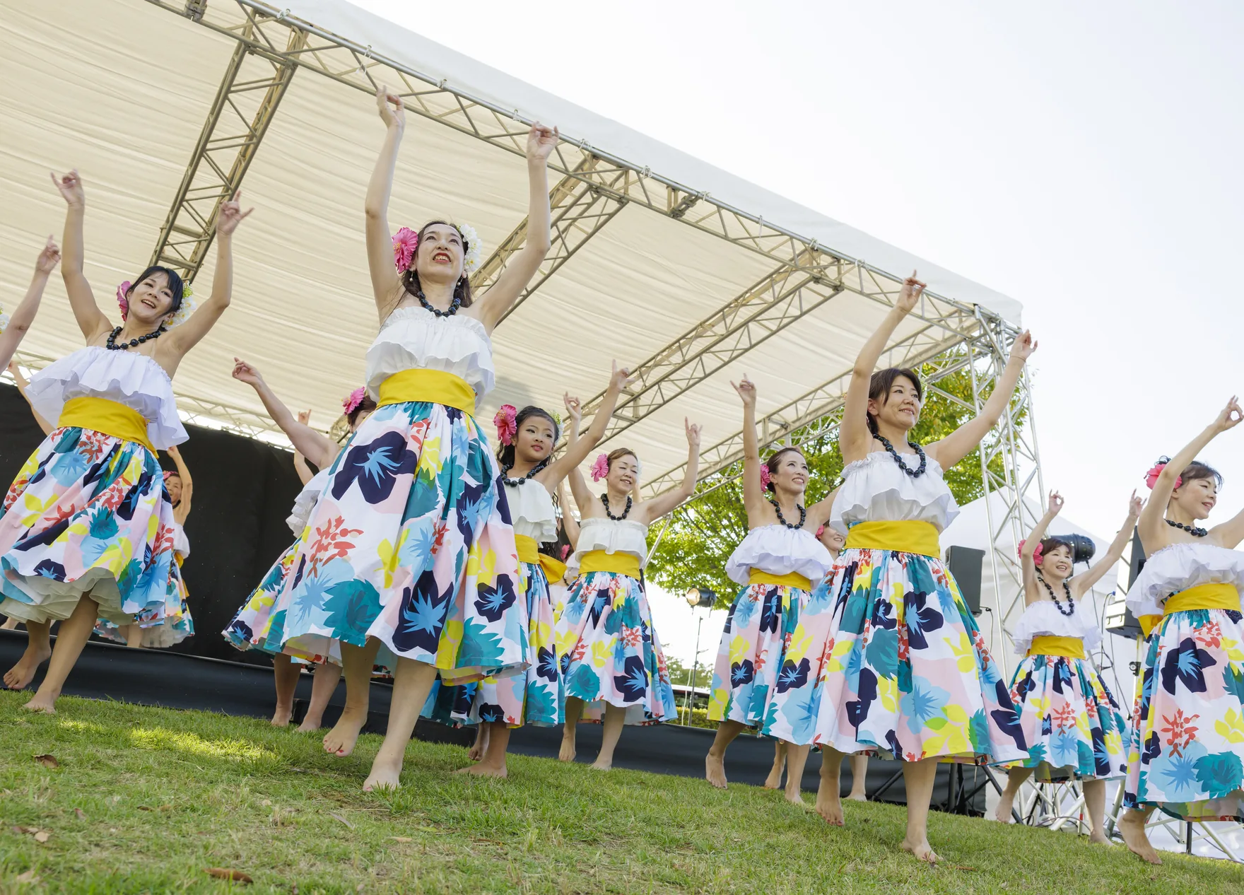 屋外ステージ前の芝生で花柄の衣装をまとってフラを踊る出演者たち